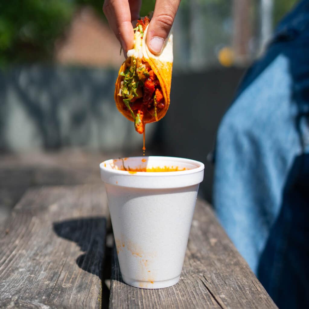 A hand dipping a birria taco into consomme from Los Agavez